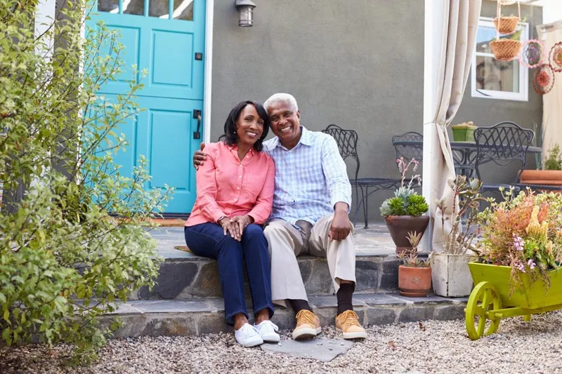 Una pareja de personas mayores sentada en su porche delantero con plantas y muebles de jardín