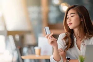 woman with long hair looking at credit card in hand at coffee shop table with laptop