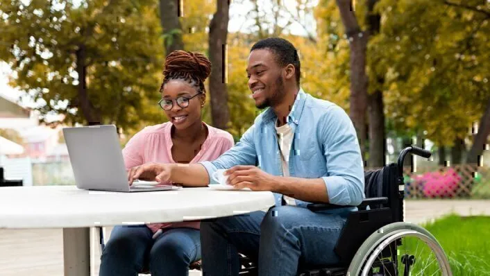 two smiling people outside at table looking at laptop, one sitting in chair, one person sitting in wheelchair