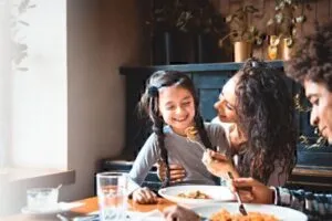 family of three eating at hoome, mother holds daughter, sharing a bite of food on fork
