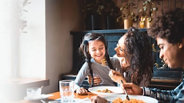 Familia de tres comiendo en casa, la madre sostiene a su hija y comparte un bocado de comida en un tenedor