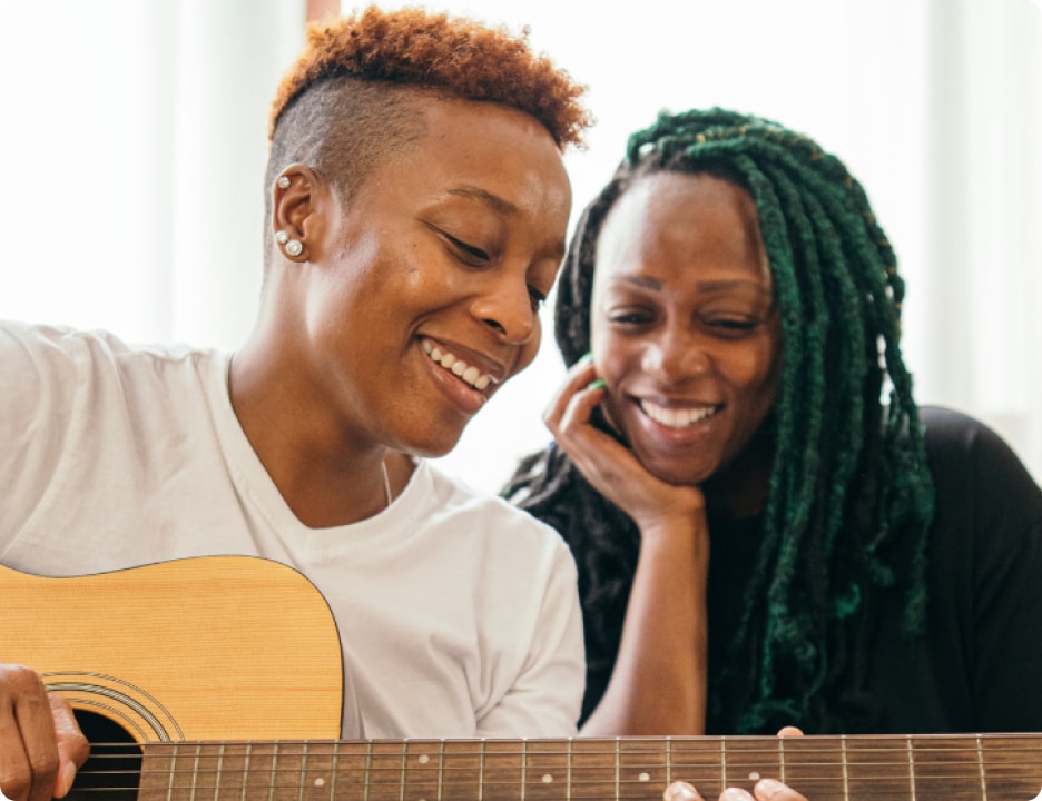 Two black women smiling. One woman is playing the guitar while the other listens.