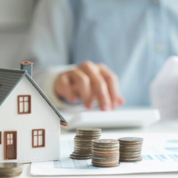 small plastic house with dark roof and red trimmed windows and stacks of coins next to the house.