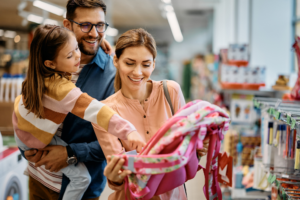 Little girl and her parents choosing backpack for school while shopping in store together.