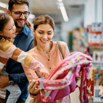 Little girl and her parents choosing backpack for school while shopping in store together.